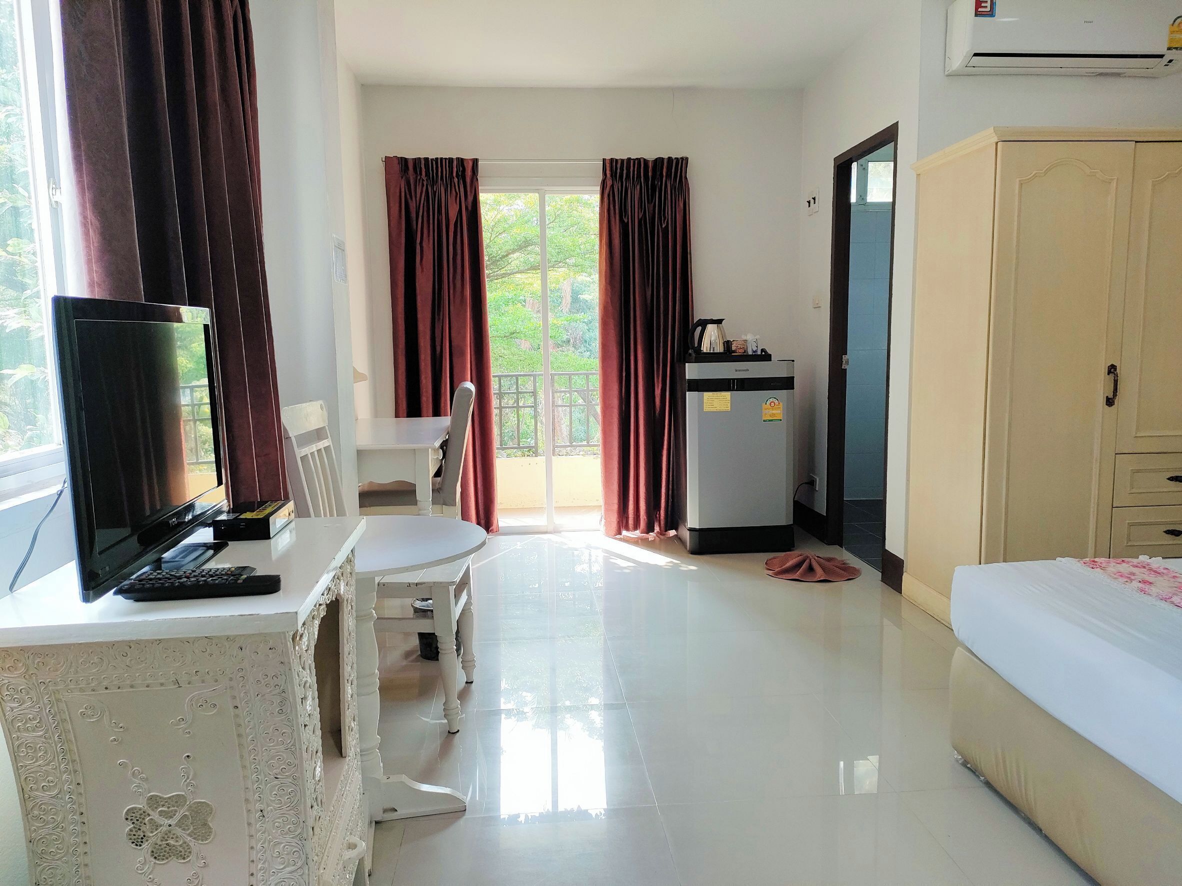 A hotel room with a bed, a desk with a computer, a chair, a kitchenette, and a view of greenery through the window.