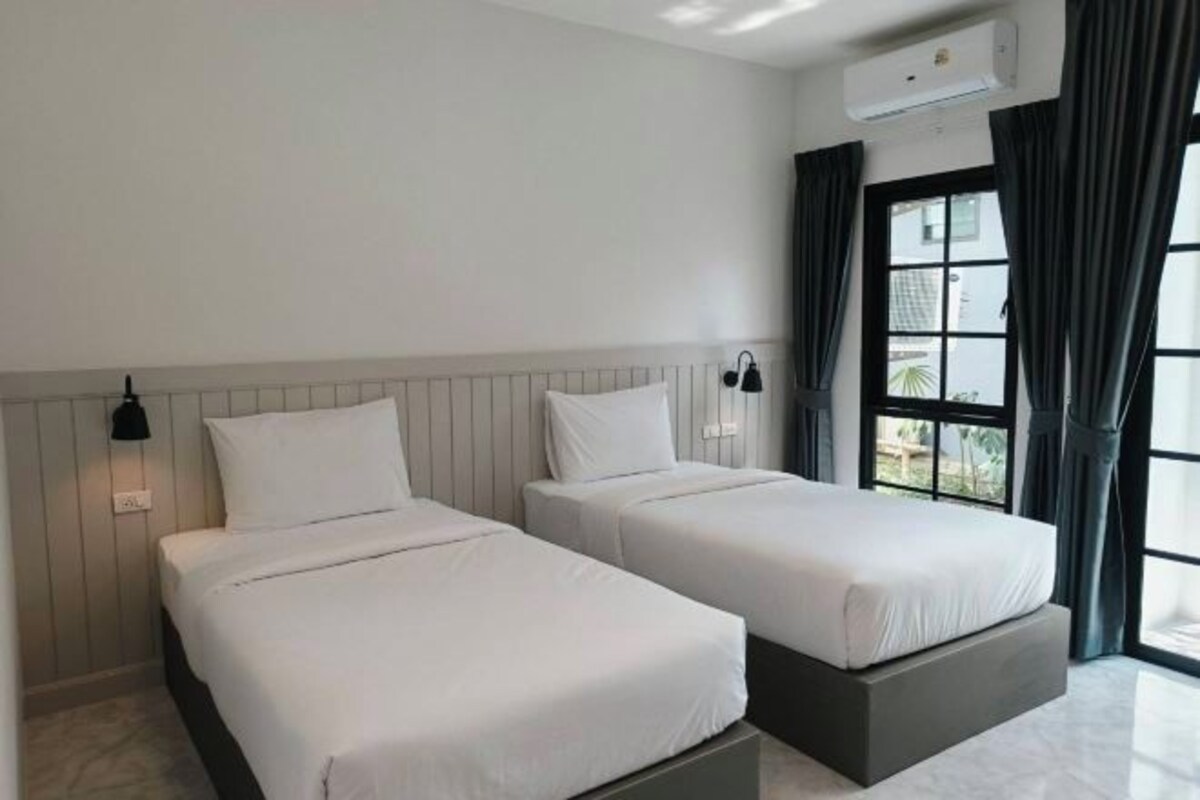 A hotel room with two beds, a window with curtains, an air conditioning unit, and a wall-mounted lamp.
