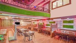 En restaurant med trægulv, flere spiseborde og stole, en balkon med gelænder og en væg med grøn kunstgræs.