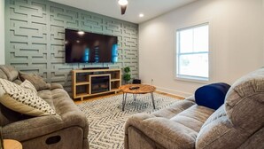 A modern living room with a ceiling fan, a flat-screen TV mounted on a geometric-patterned wall, a fireplace, and a large window providing natural light. - The Kirkwood Escape!! (Atlanta)