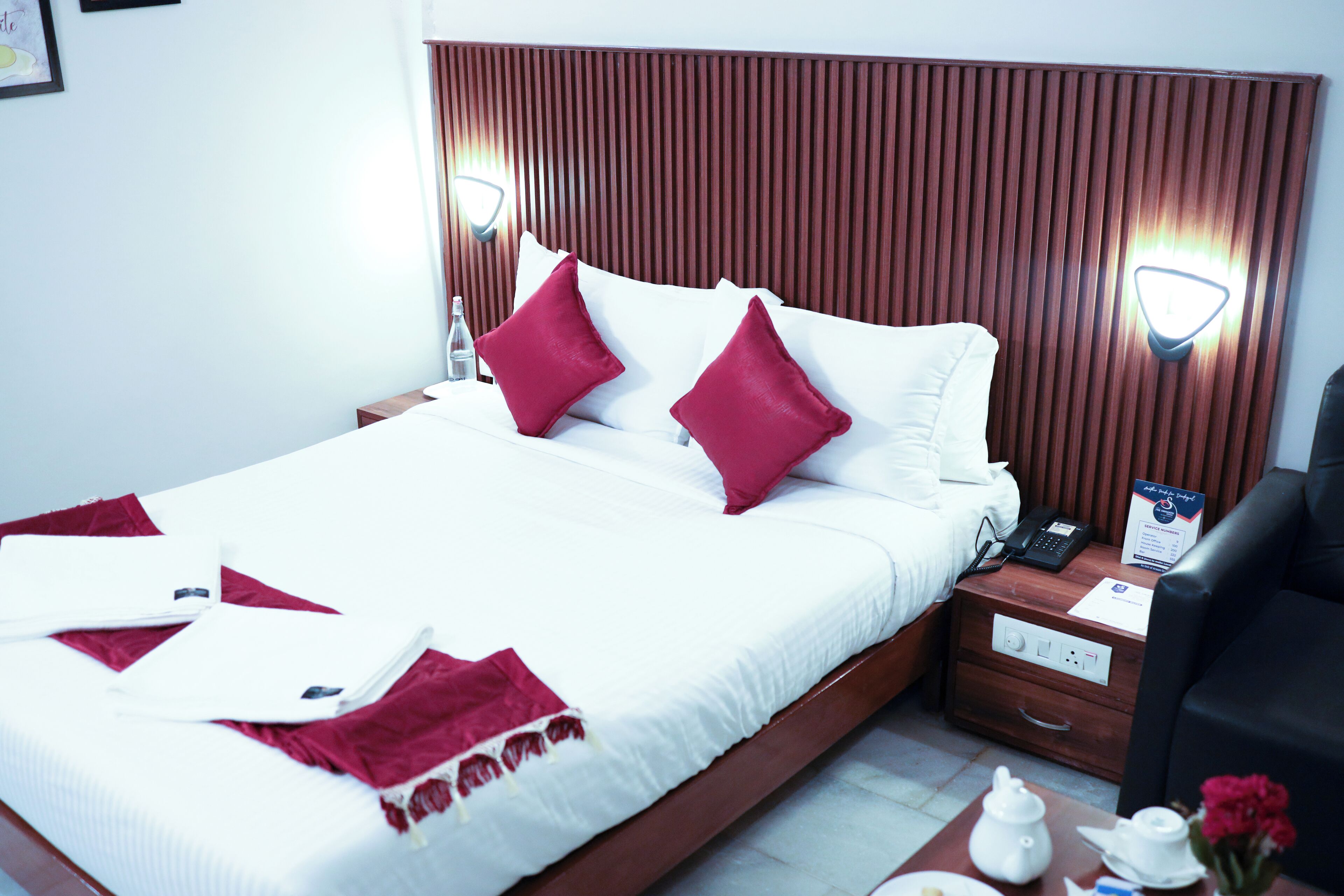 Habitación de hotel con una cama grande, dos almohadas rojas, una mesita de noche con un teléfono, una lámpara y una pequeña mesa con una tetera y flores.