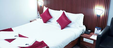 Habitación de hotel con una cama grande, dos almohadas rojas, una mesita de noche con un teléfono, una lámpara y una pequeña mesa con una tetera y flores.