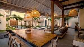 Un espace intérieur spacieux comprenant une table à manger en bois, des chaises en osier et un coin salon confortable avec des coussins.