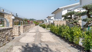 A narrow pathway flanked by stone walls and greenery, leading to modern buildings with a clear sky above.