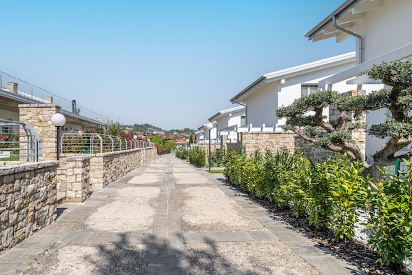 A narrow pathway flanked by stone walls and greenery, leading to a row of modern white buildings with stone accents. - 0807BS - Garda Lido Red (Padenghe sul Garda)