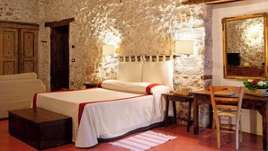 A bedroom with a stone wall, a bed with a red and white bedspread, a wooden nightstand, a desk with a chair, and a mirror on the wall.