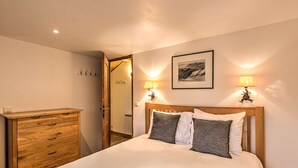 Ein Schlafzimmer mit einem Holzkoffer, einem Bett mit weißen und grauen Kissen, zwei Wandlampen und einem gerahmten Bild an der Wand.