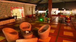 An outdoor seating area with wicker chairs and tables, a covered roof, and a checkered floor pattern.