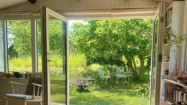 Eine offene Schiebetür führt in einen Garten mit einer Holzterrasse, einem Schaukelstuhl und einem Bücherregal.