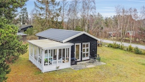 A small cabin with a covered porch and a separate glass room, surrounded by trees and a grassy yard.