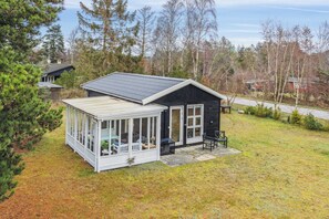 A small cabin with a covered porch and a separate glass room, surrounded by trees and a grassy yard.