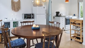 A modern kitchen with a round wooden table, chairs, a microwave, refrigerator, and a wooden shelf.