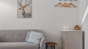 Een kamer in neutrale tinten met een grijze bank, een klein krukje, een kastje met kaarsen en een houten rek voor kleding.