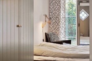 A bedroom with a bed, a nightstand, a lamp, a window with a view of greenery, and a door with a frosted glass panel.