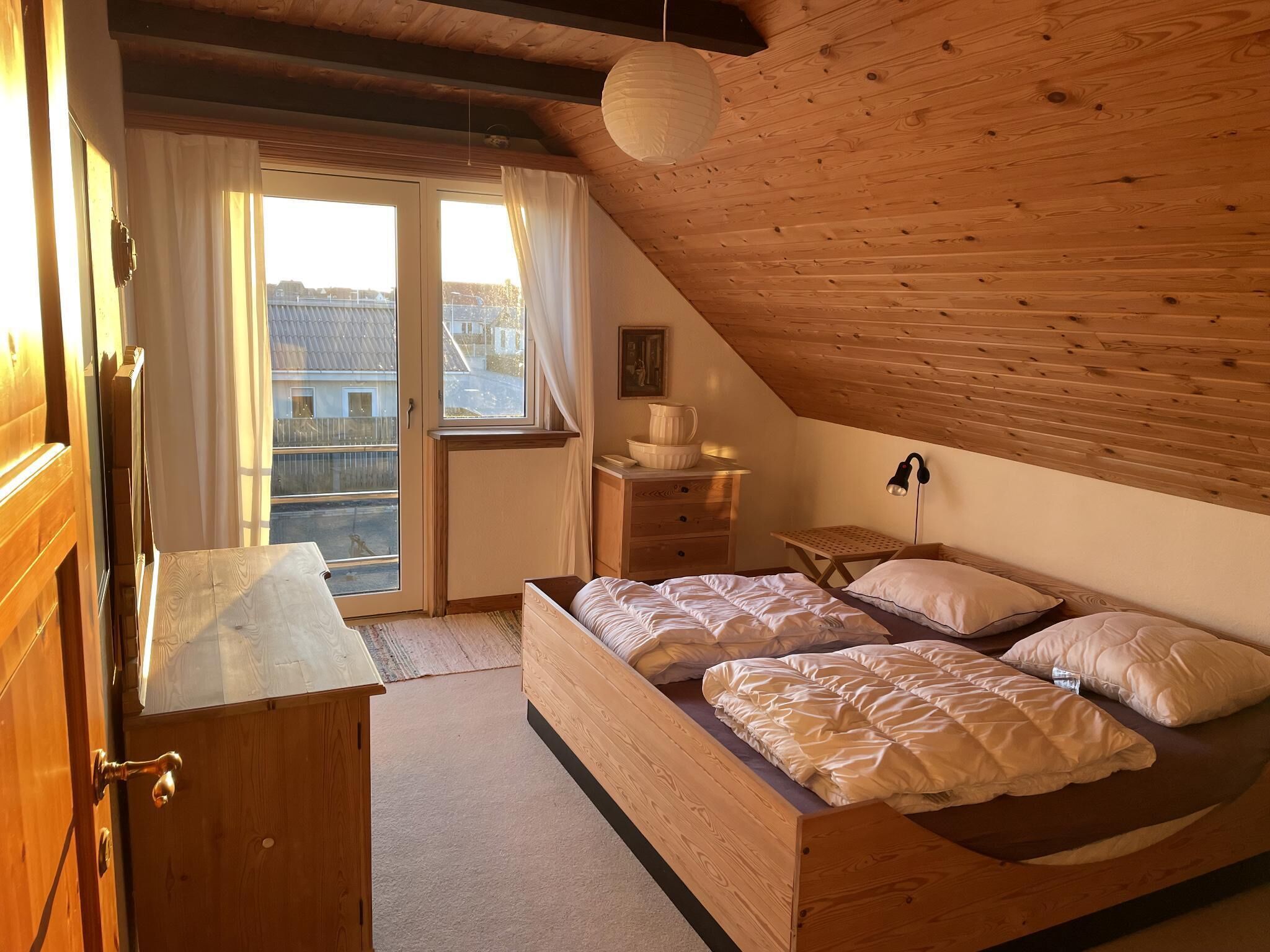 A bedroom with a wooden ceiling, a bed with white bedding, a nightstand, a lamp, and a view of the outside through a window.