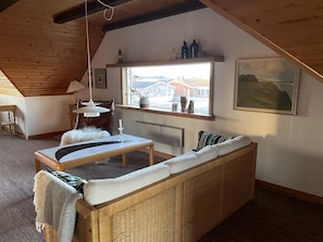 Een gezellige kamer met een hoge plafond, een rieten zithoek, een kleine tafel, een raam met uitzicht en een ingelijst beeld aan de muur.