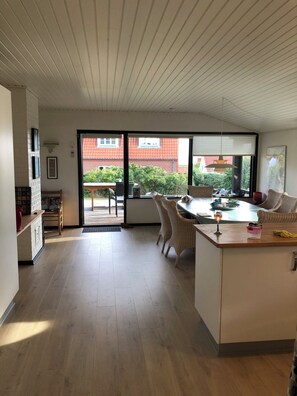 A modern kitchen with a large island, a dining area with wicker chairs, and a view of a red-roofed building through large windows.
