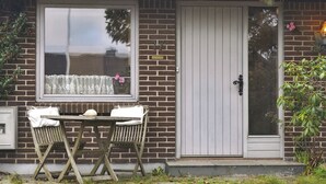 Una casa de ladrillo con puerta blanca, una ventana con cortinas de encaje y una pequeña mesa con sillas afuera.