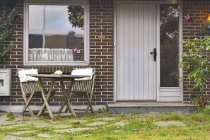 A brick house with a white door, a window with lace curtains, and a small table with chairs outside.