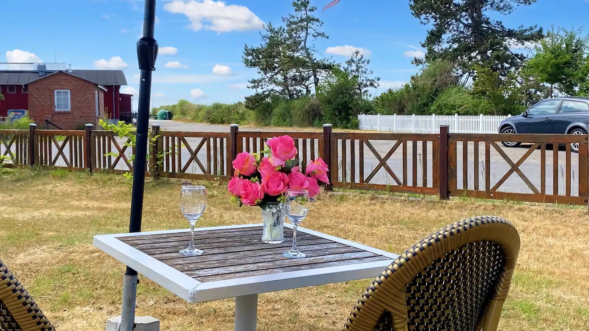 Une table en bois, avec deux verres à vin et un vase de fleurs roses, est placée à l’extérieur, avec une clôture en bois et une voiture en arrière-plan.
