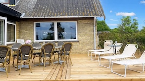 Een houten terras met buitenmeubilair, een stenen gebouw met grote ramen, en een heldere lucht.