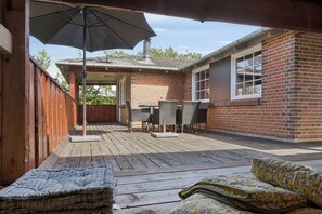 A covered patio area with a table and chairs, a wooden deck, and a brick building.