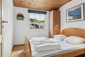 Un dormitorio con cama de madera, ropa de cama blanca, una ventana con vista a una casa y un cuadro en la pared.