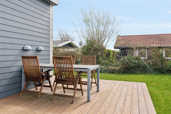 Eine Holzterrasse mit Tisch und Stühlen, ein graues Gebäude mit Satellitenschüssel und ein Garten mit Hecke und Bäumen.