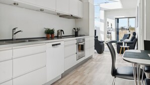Eine moderne Küche mit weißen Schränken, einer Edelstahlspüle und einem eingebauten Backofen. Ein Essbereich mit Tisch und Stühlen bietet einen Blick ins Freie durch große Fenster.