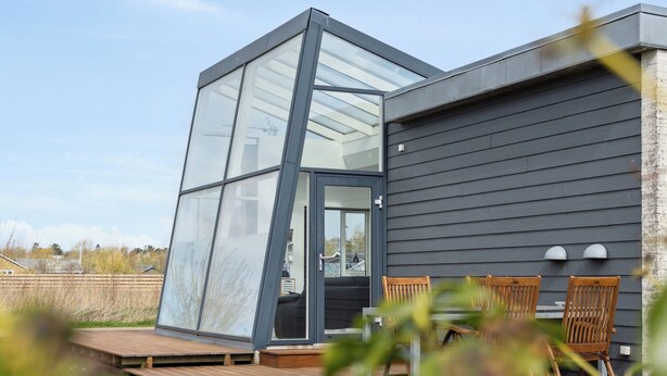 Ein modernes Haus mit einer großen Glasveranda, einer Holzterrasse und Sitzgelegenheiten im Freien.