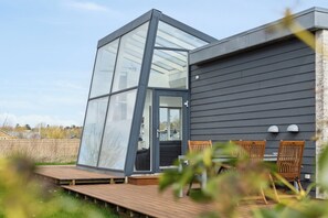 Una casa moderna con un amplio ventanal de vidrio, terraza de madera y mobiliario exterior.