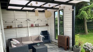 An outdoor patio with a sofa, tables, hanging lights, and potted plants. - Cozy cottage close to the beach. (Bogense)