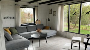 A modern living room with a grey sectional sofa, a round coffee table, and a view of the outdoors through large windows. - Cozy cottage close to the beach. (Bogense)