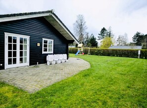 A black building with a white door and windows, a paved area with benches, and a grassy yard with a swing set.