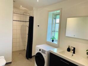 A bathroom with a shower, a toilet, a washing machine, a sink, and a window with a view.