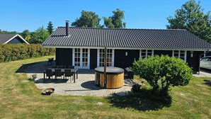 Una casa a due piani con tetto in tegole, un portico con tavolo e sedie, e un piccolo giardino con una botte di legno e un cespuglio.