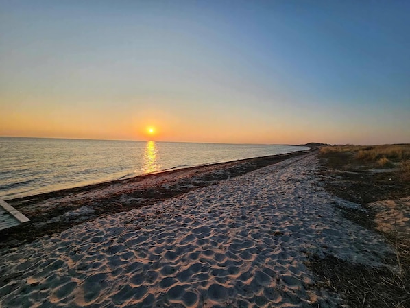A beach with a wooden walkway leading to the water, a sunset in the background, and a clear sky.