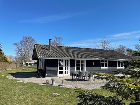 A single-story building with a dark roof and white trim, a patio with outdoor furniture, and a small garden area.