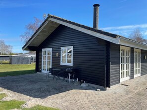 A black wooden building with white-framed windows, a chimney, and a patio area with outdoor seating. - Lovely cottage with secluded garden (Rødby)