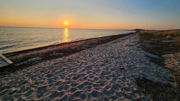 Una spiaggia con una passerella di legno che conduce all'acqua, un tramonto sullo sfondo e un cielo sereno.