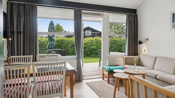 A room with a sofa, chairs, a table, and a view of a garden through large windows.