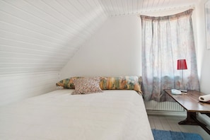 Ein Schlafzimmer mit schrägen Wänden, einem Bett mit gemusterten Bettbezügen, einem Nachttisch mit Lampe und einem Fenster mit Vorhängen.