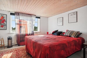 A bedroom with a wooden ceiling, a bed with a red patterned bedspread, a window with a view of a red telephone booth, and a framed picture on the wall.