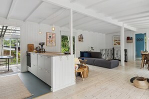 A spacious living area with a grey sofa, a kitchen with white cabinetry, and a dining area with a wooden table.