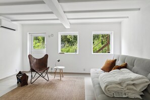 Ein Zimmer mit einer Couch, einem Sessel, einem kleinen Tisch und zwei Fenstern mit Blick auf Grünflächen.
