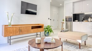 A modern living room with a wooden cabinet, a round coffee table, a sofa, a TV mounted on the wall, and a kitchen area in the background. - Beco Santa Emilia 2I, a Home in Madeira (Funchal)