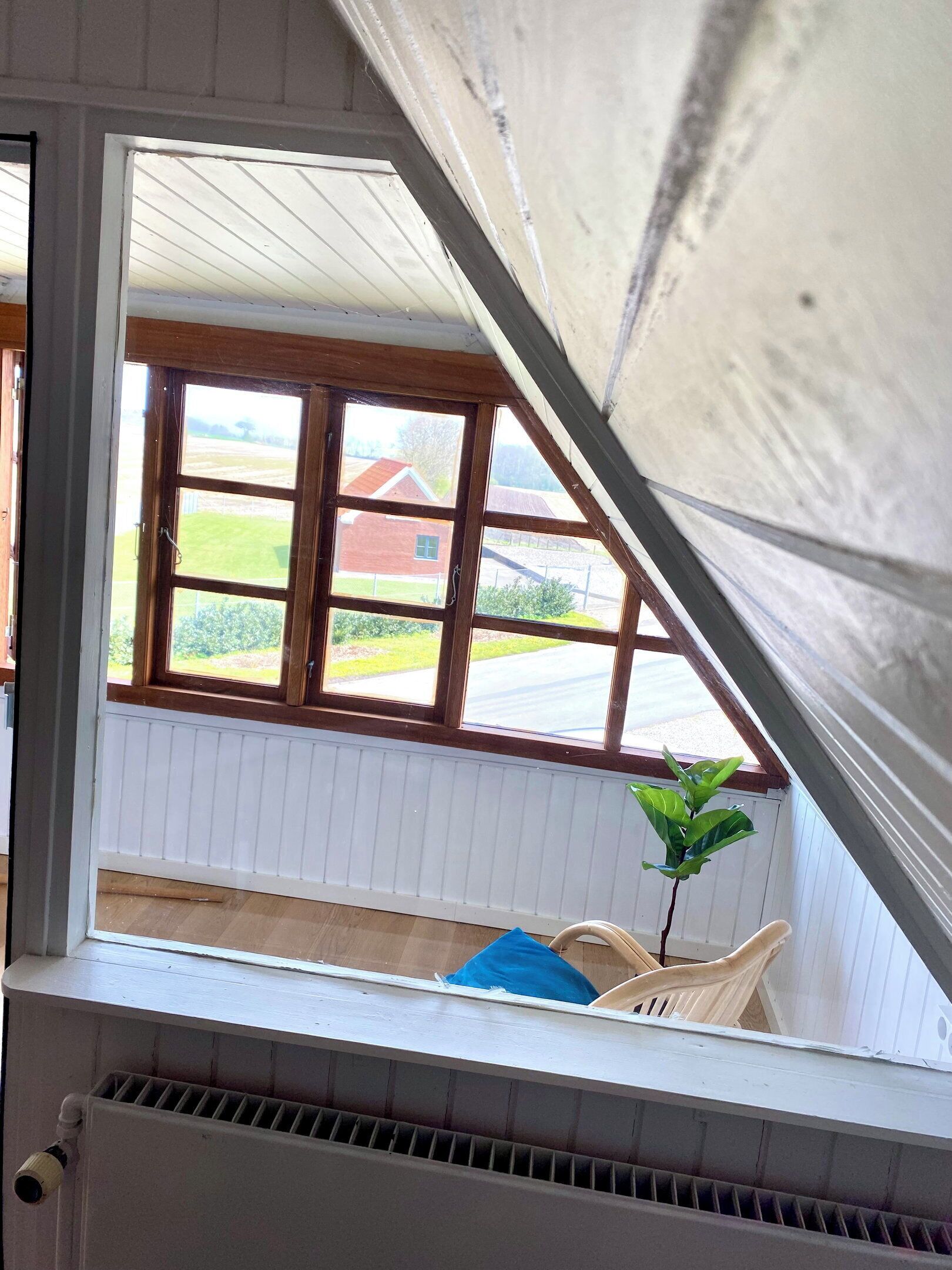Une pièce avec un plafond en pente, une fenêtre donnant sur un bâtiment rouge et sur de la verdure, un radiateur et une chaise.