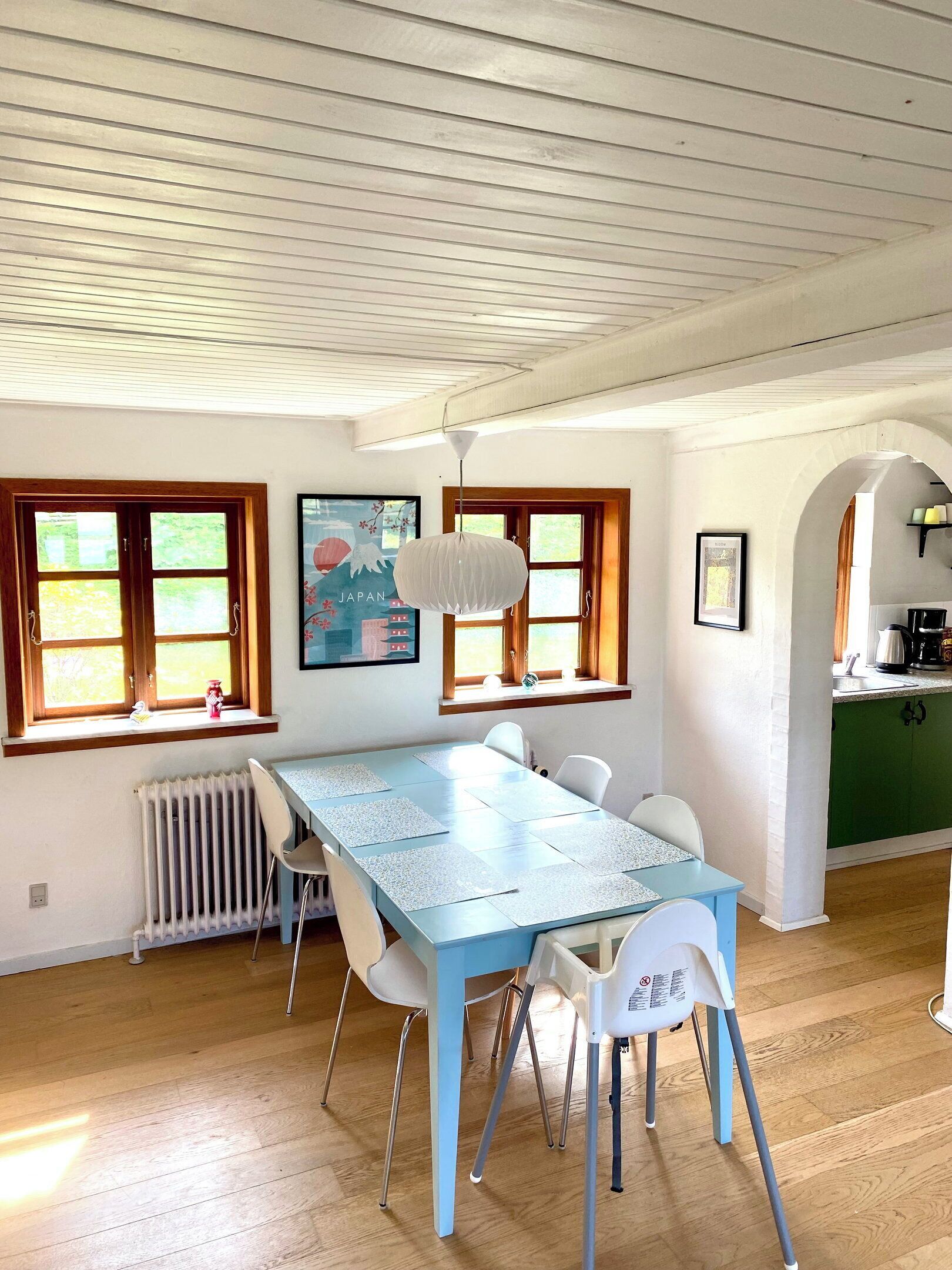 A dining area with a blue table, white chairs, a hanging light fixture, and a kitchen visible through an archway.