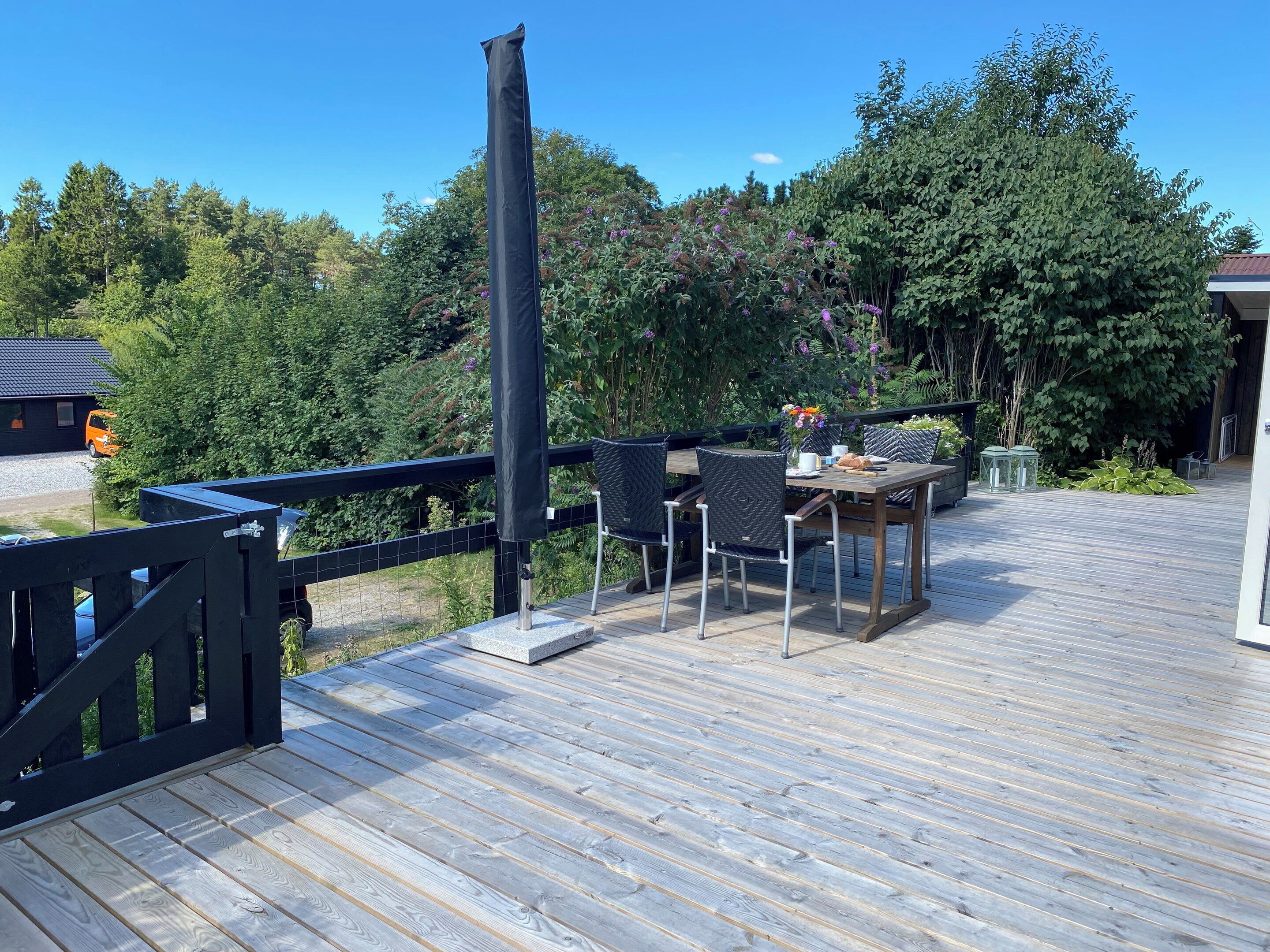 Una terraza de madera con una mesa y sillas, rodeada de follaje y una cerca negra.