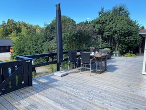 A wooden deck with a table and chairs, surrounded by greenery and a black fence.